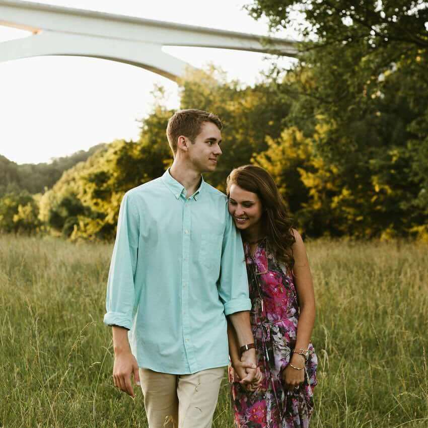 A young couple holds hands in a grassy field with a bridge in the background in Williamson County, Tennessee.