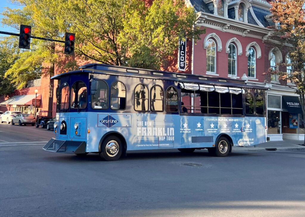 A blue trolley bus with 'Franklin Hop-On Hop-Off Tour' signage travels down a city street in Williamson County, Tennessee.