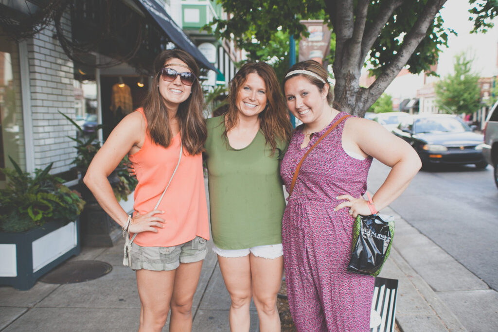 Three women in summer dresses stand arm-in-arm on a tree-lined sidewalk, smiling in Williamson County, Tennessee.