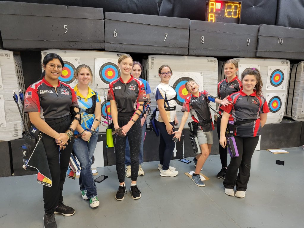 Archery Summer Camp A group of eight young women, dressed in matching archery uniforms, pose together in front of a wall of archery targets in an indoor range in Williamson County, Tennessee.