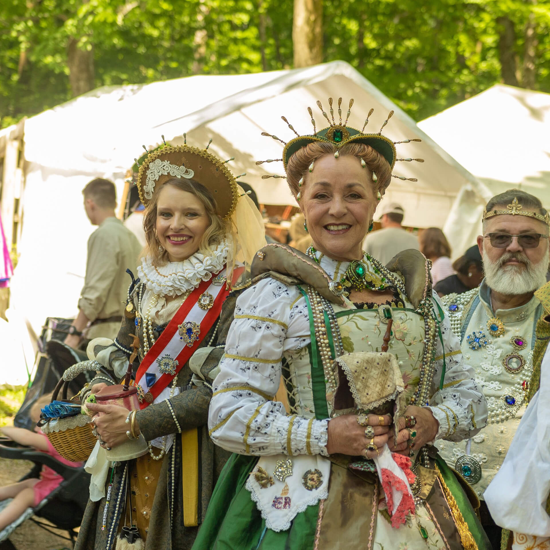 Two women dressed in elaborate Renaissance-era costumes, adorned with jewels and crowns, stand smiling amidst a festive outdoor gathering with white tents and trees in the background in Williamson County, Tennessee.