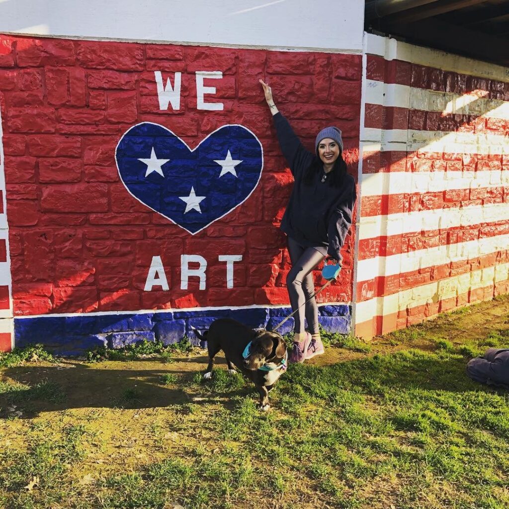 Woman stands beside a colorful mural with a dog on a leash, smiling in Williamson County, Tennessee.