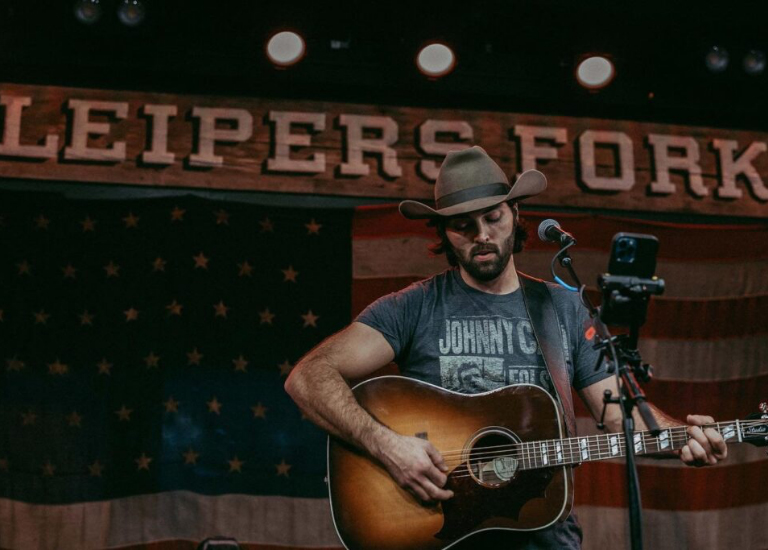A musician wearing a cowboy hat plays an acoustic guitar on stage with an American flag backdrop in Williamson County, Tennessee.