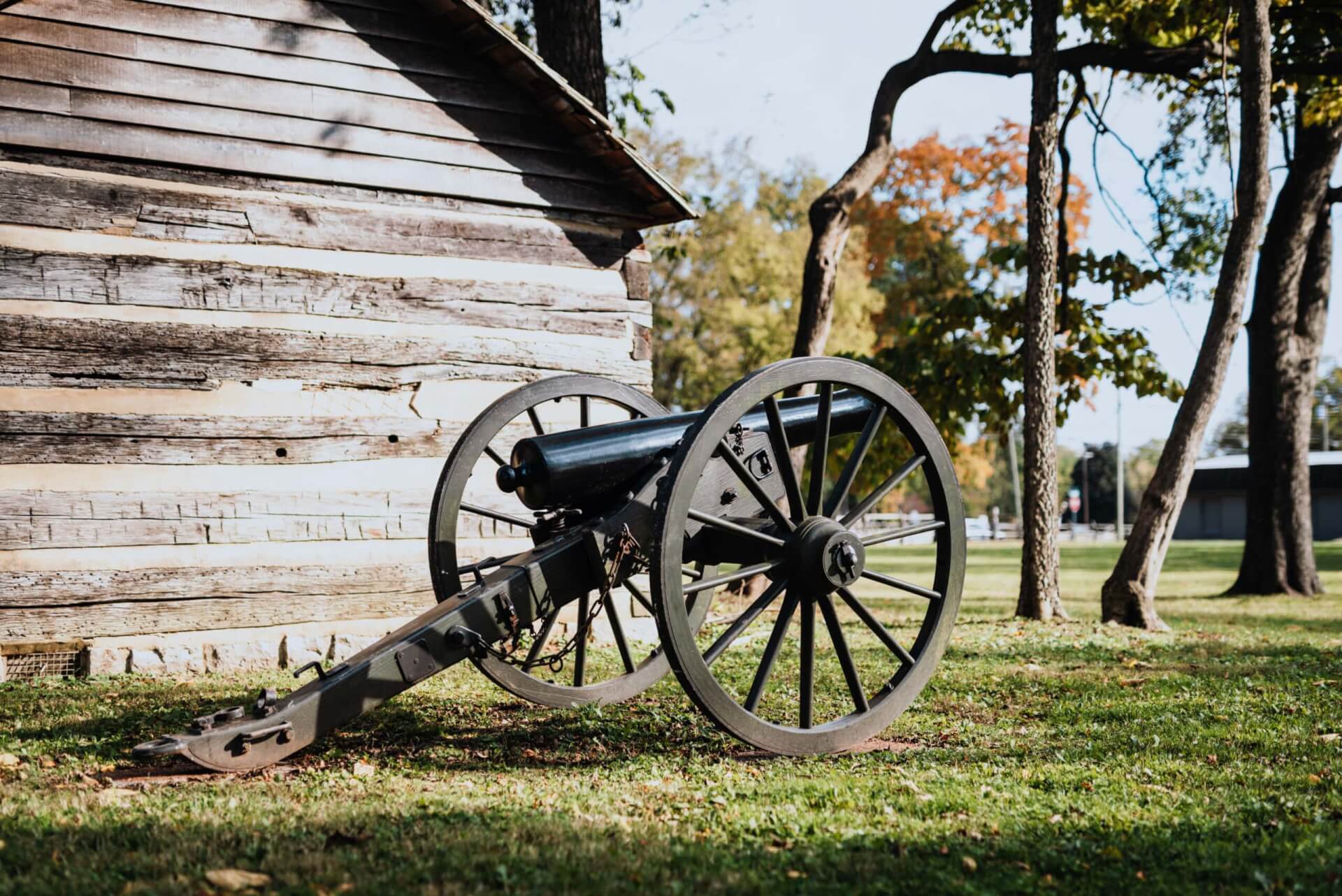 An old cannon with large wheels rests on grass beside a rustic wooden cabin in Williamson County, Tennessee.