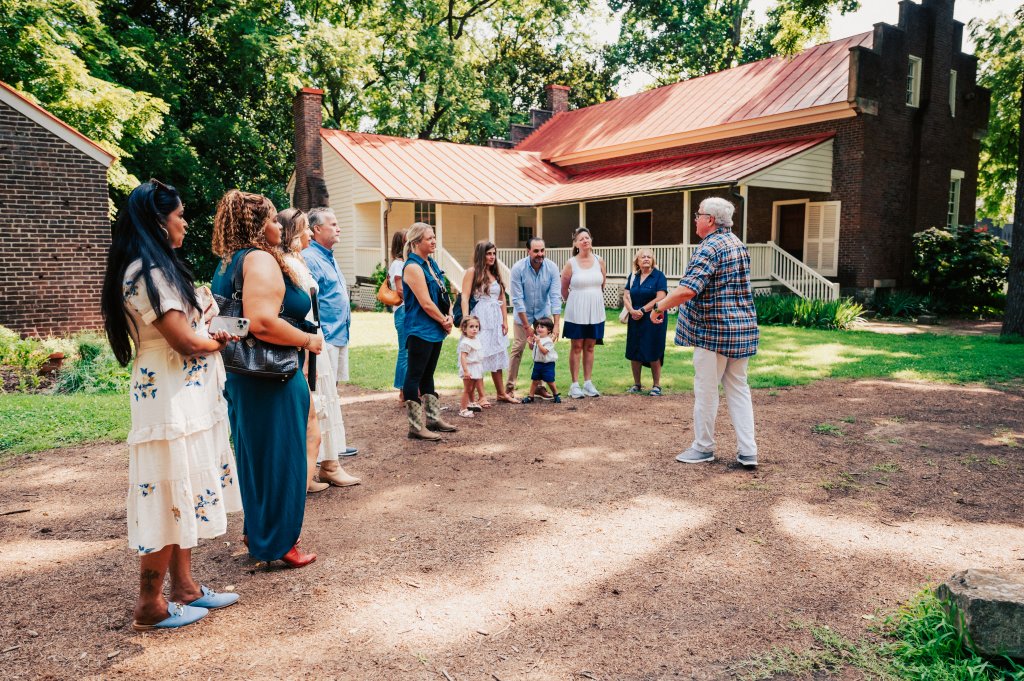 Franklin, Tennessee's Preservation Success Story A diverse group of people, including adults and children, gather in a circle on a dirt path in front of a historic brick house with a red roof, surrounded by lush greenery and gardens in Williamson County, Tennessee.