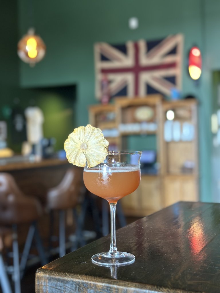 A cocktail with a dried pineapple garnish sits on a wooden bar, with a Union Jack in the background in Williamson County, Tennessee.