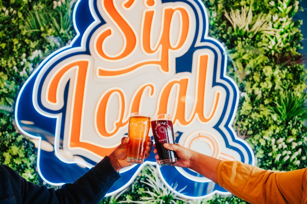 Kick Back at These 6 Action-Packed Spots for Fun & Games in Franklin Two people clinking glasses of beer in front of a 'Sip Local' neon sign in Williamson County, Tennessee.