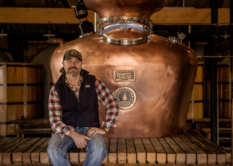 Man sits on wooden platform next to large copper still in industrial setting in Williamson County, Tennessee.