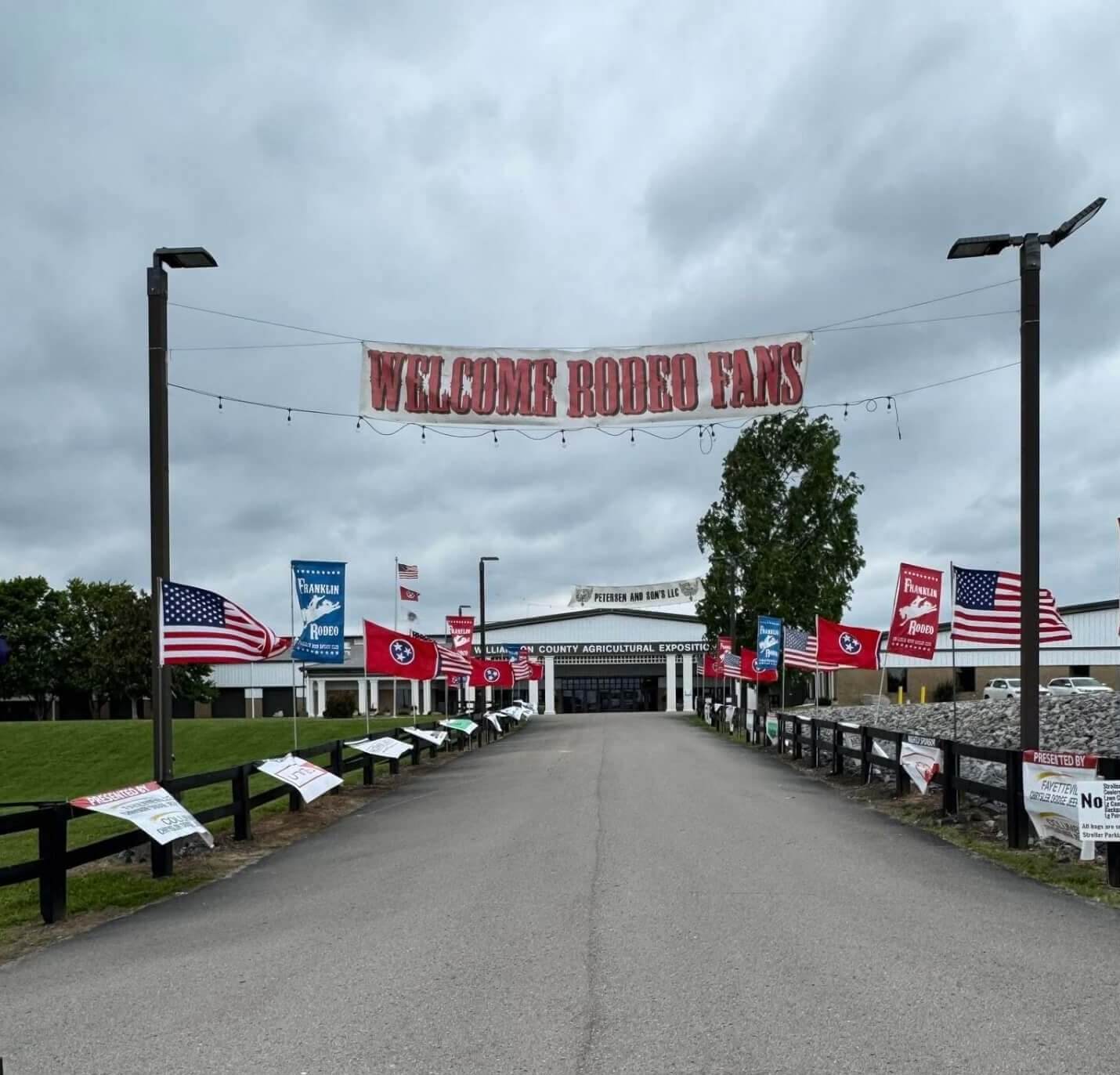 A wide, empty road leads towards a large building adorned with numerous flags, including American and rodeo-themed banners, under a cloudy sky in Williamson County, Tennessee.