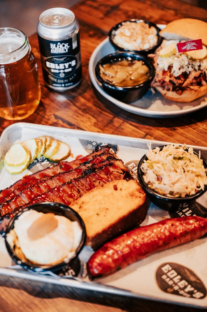 A hearty barbecue platter featuring smoked ribs, sausage, baked beans, coleslaw, and cornbread, accompanied by a can of Golden Ale and a glass of beer in Williamson County, Tennessee.