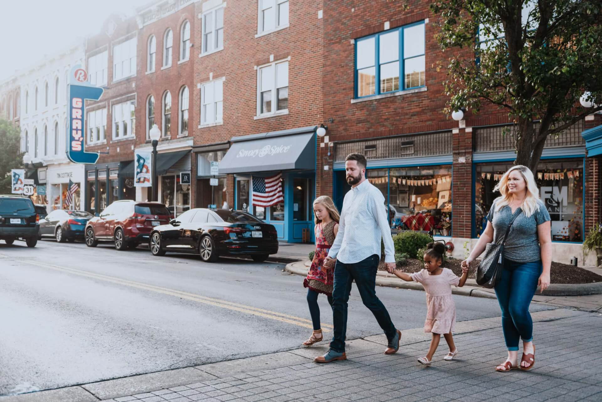 A family of four walks hand-in-hand down a quaint town street lined with parked cars and brick buildings in Williamson County, Tennessee.