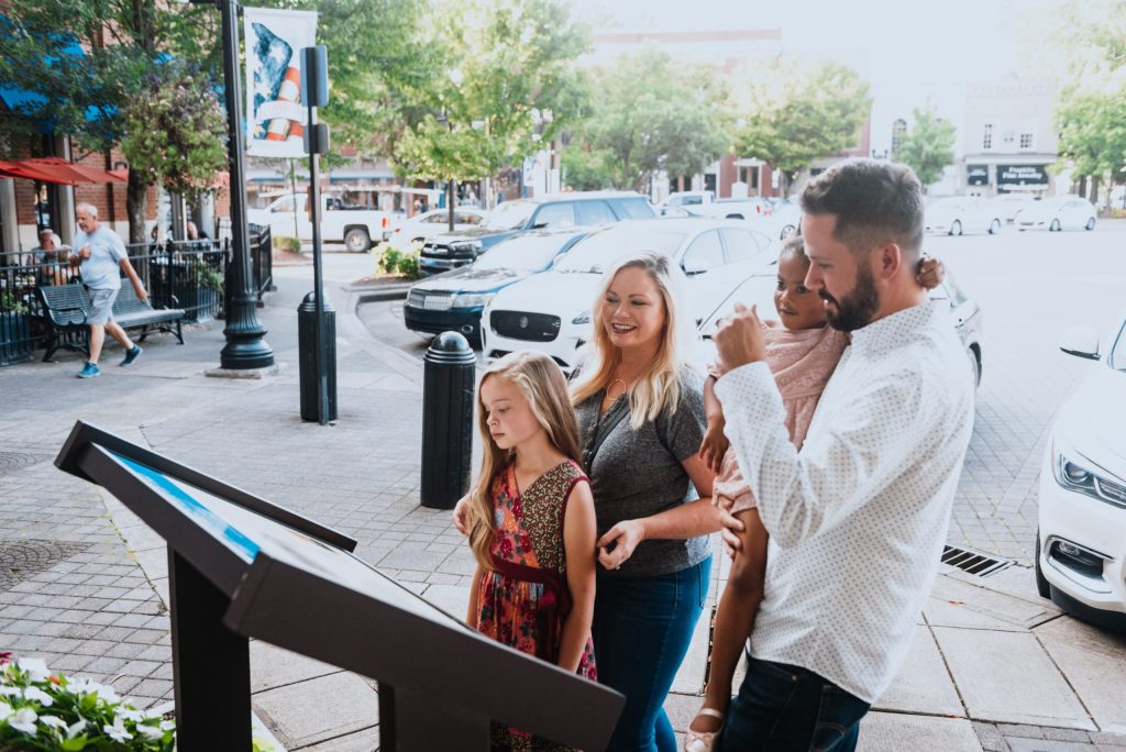 A family of four, including two young girls, gathers around an interactive display in a sunny town square in Williamson County, Tennessee.
