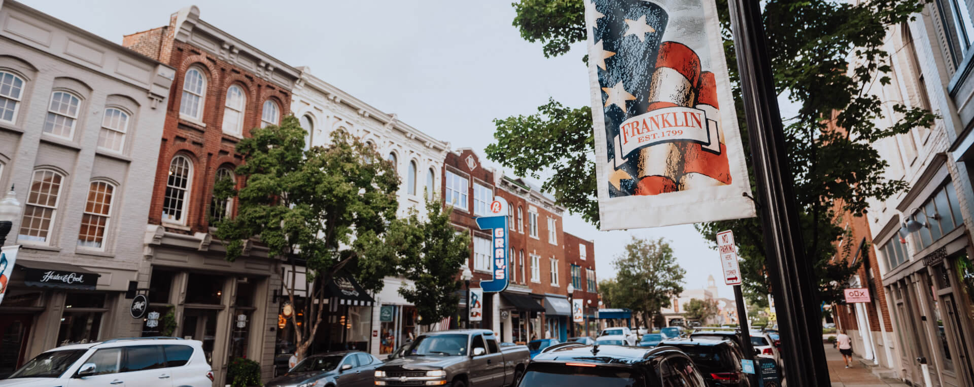 Things To Do A bustling downtown street with historic buildings, parked cars, and a patriotic banner in Williamson County, Tennessee.