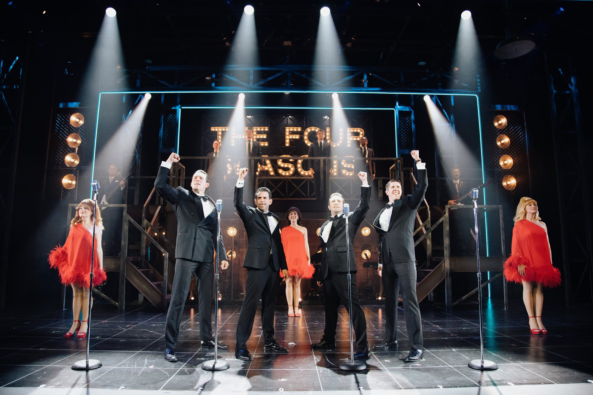 A group of five performers, dressed in elegant black tuxedos and vibrant red dresses, stand triumphantly on stage with their arms raised, illuminated by dramatic stage lights in Williamson County, Tennessee.