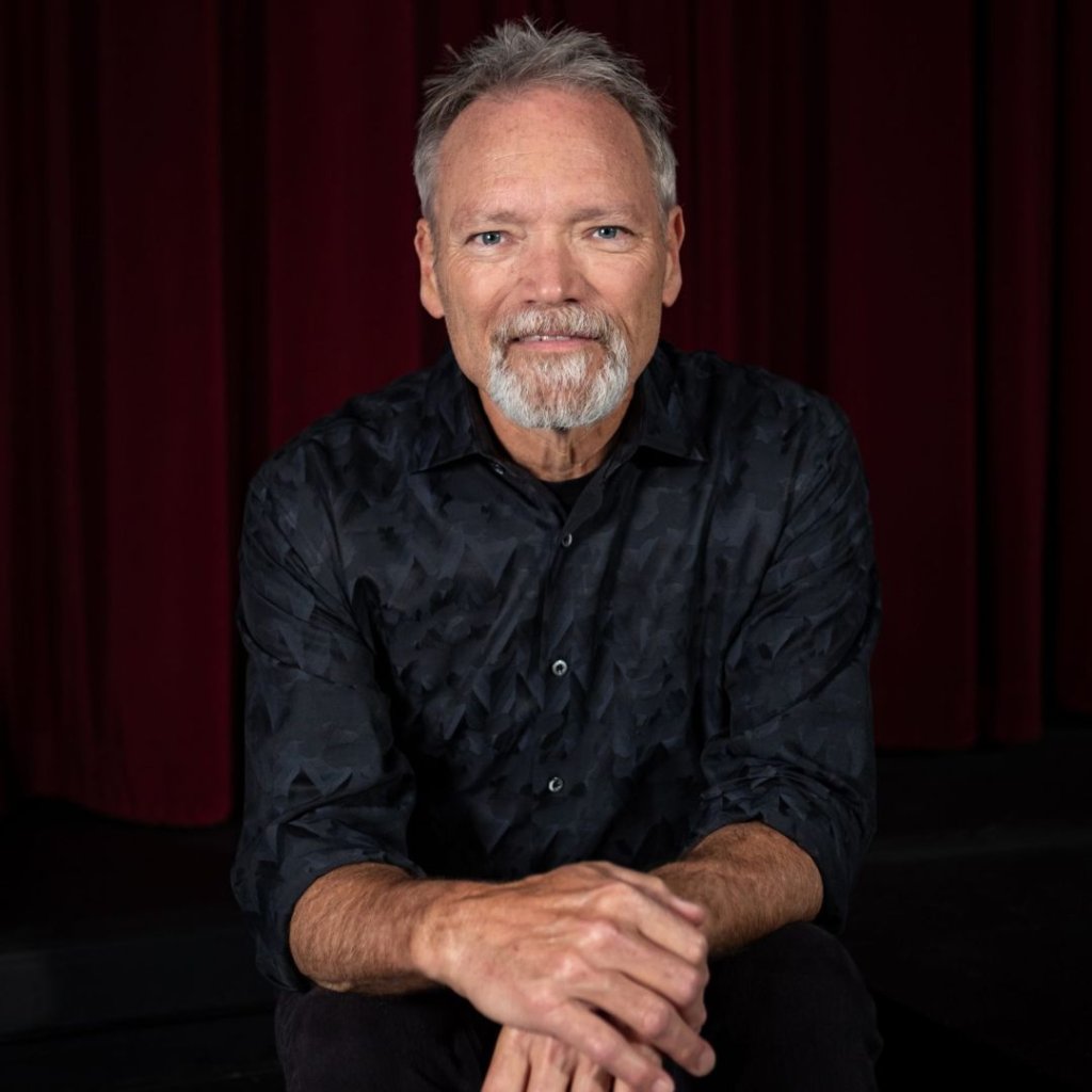 A man with gray hair and a beard, wearing a black button-up shirt with a geometric pattern, sits with his hands clasped in front of him against a dark red curtain backdrop in Williamson County, Tennessee.