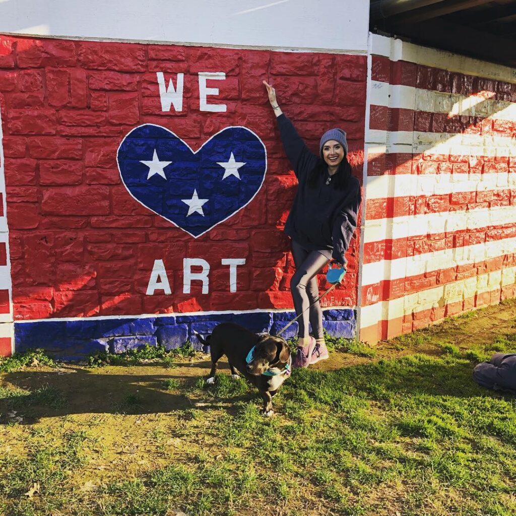 Woman poses with dog in front of patriotic mural with 'WE ART' text and heart in Williamson County, Tennessee.