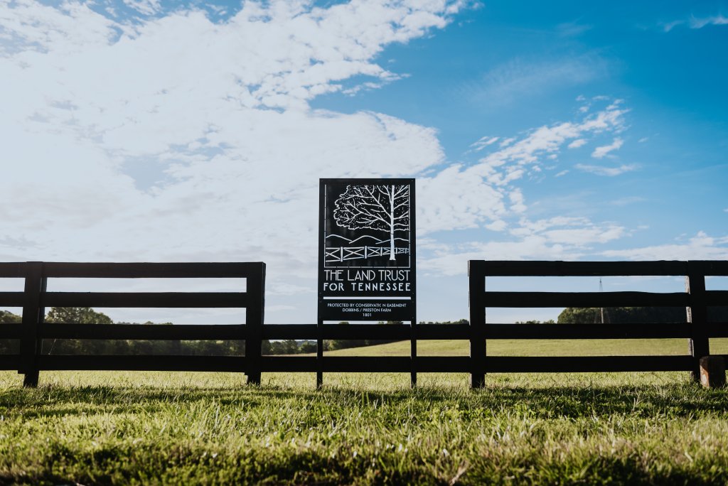Franklin, Tennessee's Preservation Success Story A wooden fence with a sign for The Land Trust for Tennessee in a grassy field under a blue sky with clouds in Williamson County, Tennessee.