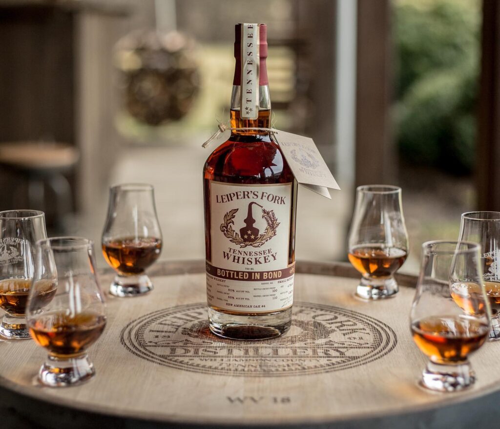 A bottle of Leiper's Fork Tennessee whiskey sits on a wooden barrel surrounded by small glasses filled with amber liquid in Williamson County, Tennessee.