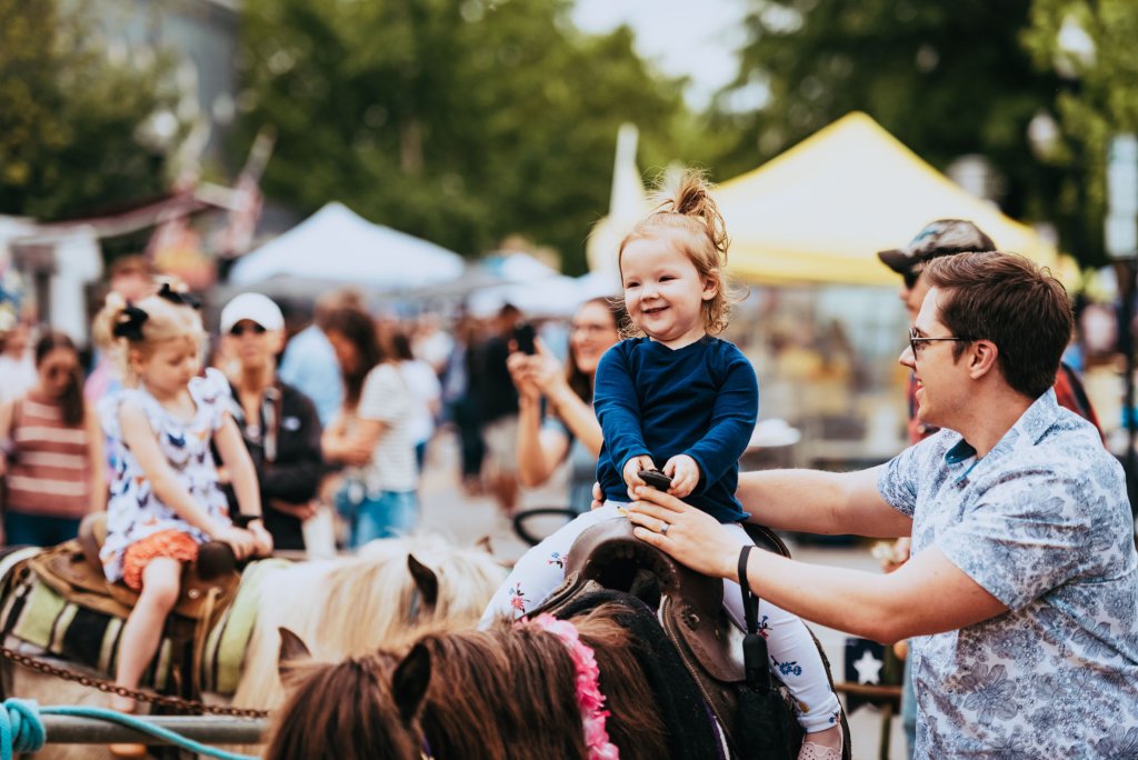 Main Street Festival A young girl in a blue shirt rides a pony at a lively outdoor festival, smiling broadly as her father guides the horse in Williamson County, Tennessee.