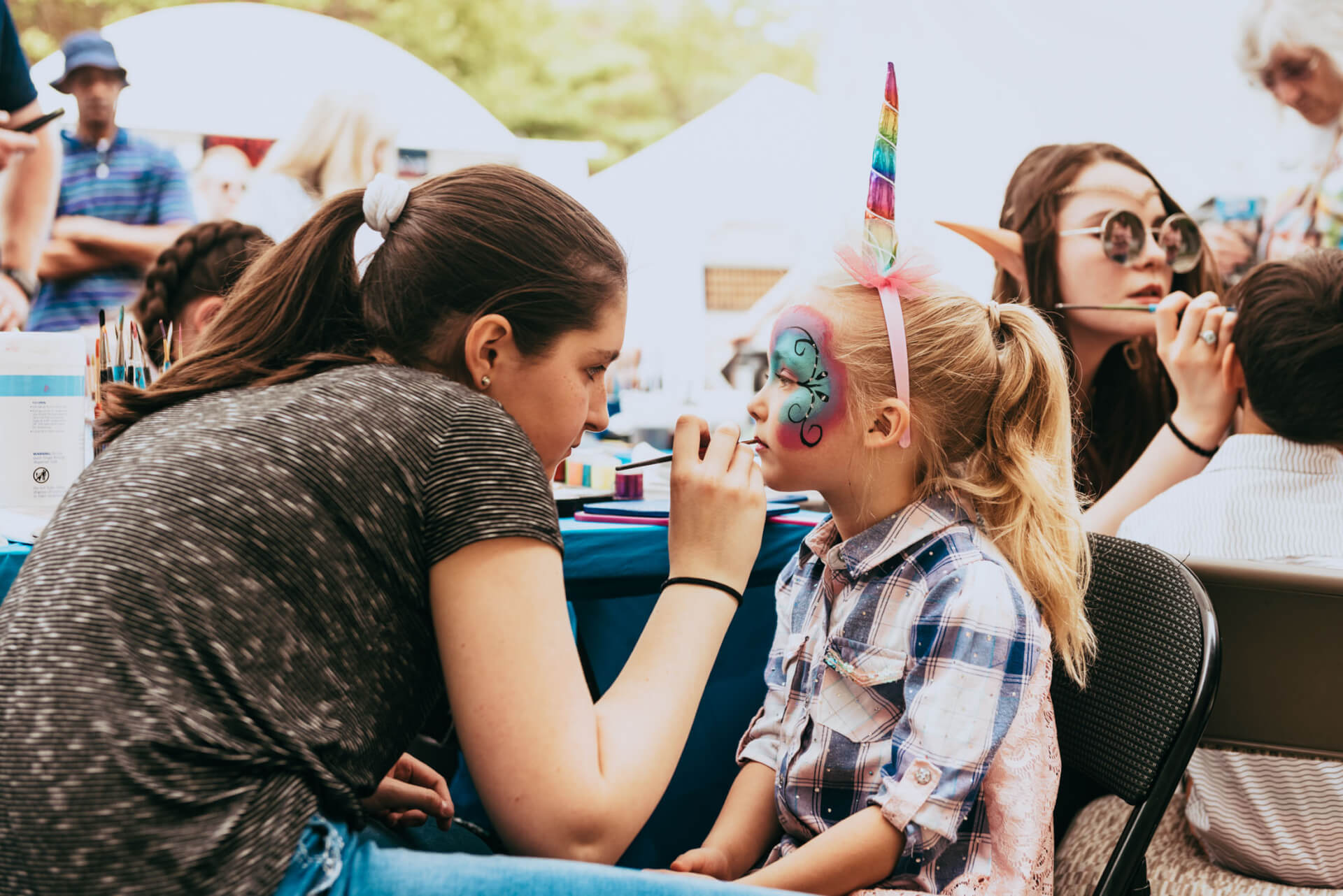 Main Street Festival A woman carefully paints a young girl's face with intricate designs at a face painting booth, while the girl wears a colorful unicorn horn headband in Williamson County, Tennessee.