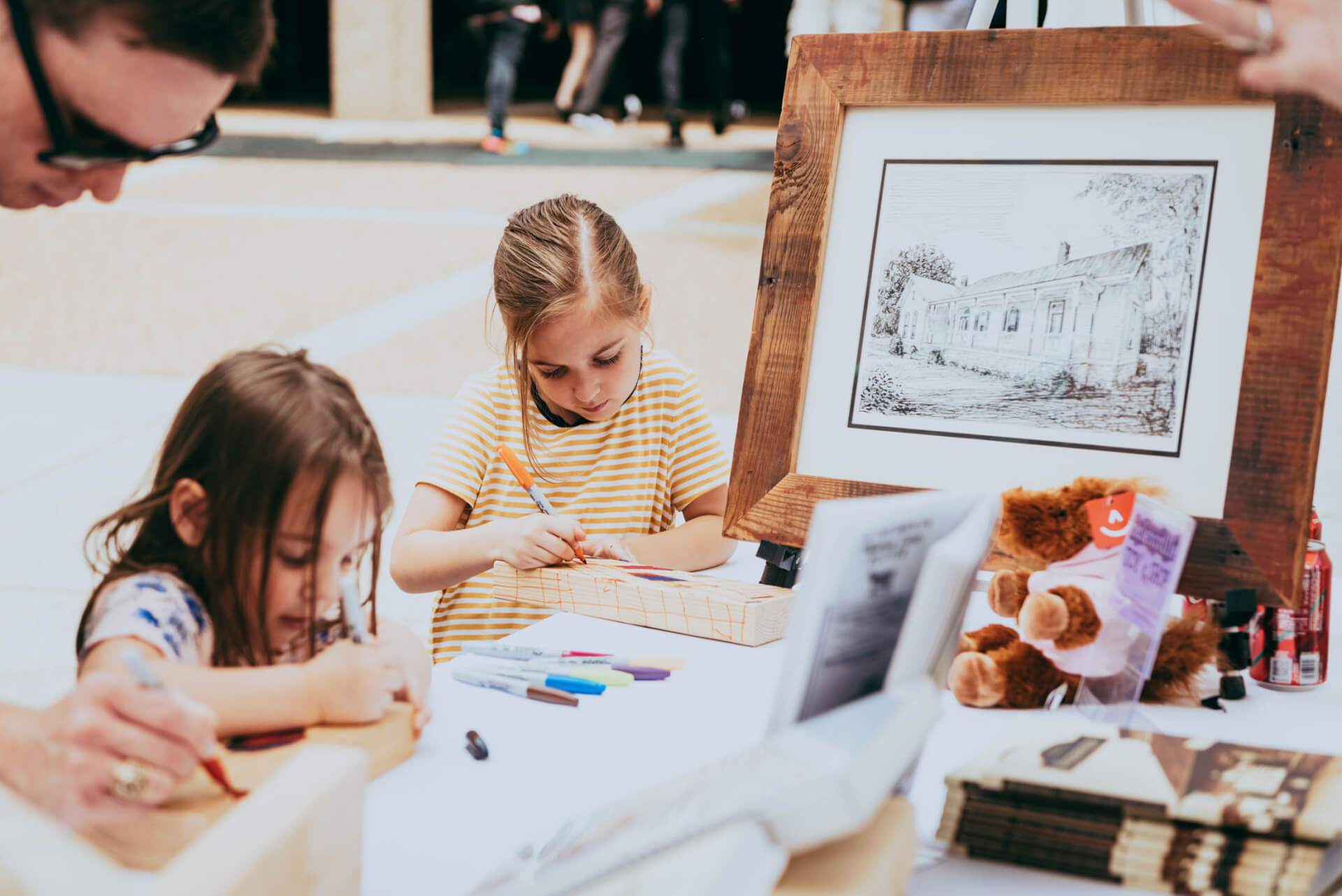 Main Street Festival Two young girls are engrossed in drawing at a table, surrounded by art supplies and a framed picture of a house, while an adult assists them in Williamson County, Tennessee.