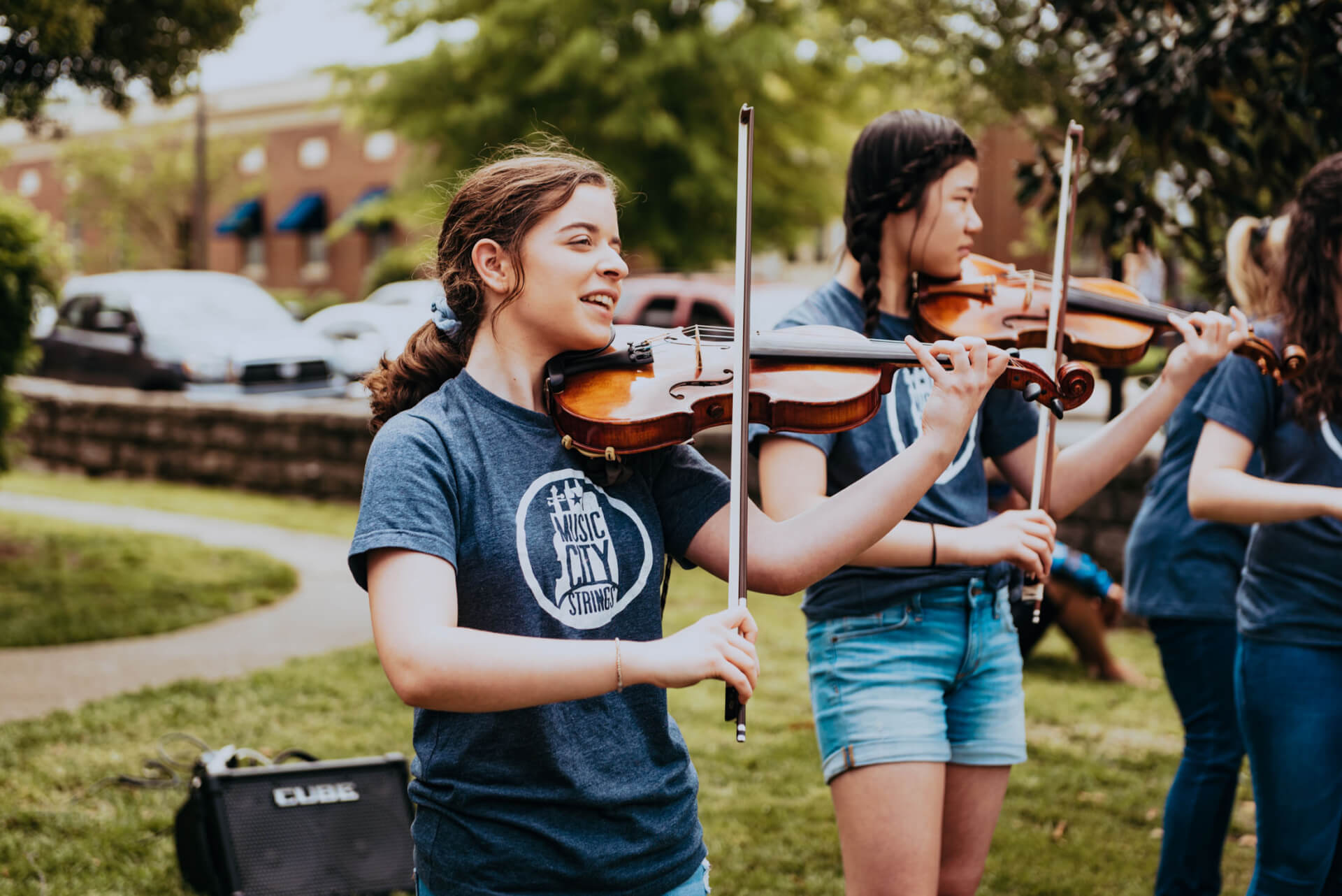 Main Street Festival A group of young women, dressed in matching blue shirts, play violins together in a park setting, with trees and a building in the background in Williamson County, Tennessee.