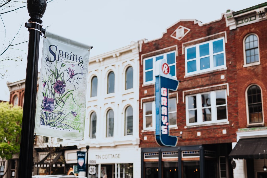 The Ultimate Guide to a Superb Spring Break in Franklin, Tennessee A charming downtown street scene with a 'Spring' banner featuring purple flowers hanging from a lamppost, set against a backdrop of historic brick and white buildings with large windows and vintage signage in Williamson County, Tennessee.