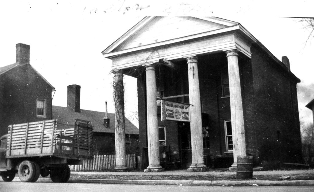 Franklin, Tennessee's Preservation Success Story A vintage black and white photograph of a two-story brick building with four tall columns supporting a triangular pediment, situated on a street corner with a wooden truck parked in front and a residential house to the left in Williamson County, Tennessee.