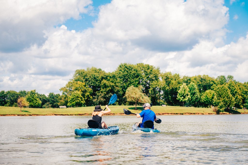 Discover 12 Fun Things To Do With Young Kids in Franklin, TN Two kayakers paddle across a serene lake surrounded by lush green trees and a grassy shore under a partly cloudy sky in Williamson County, Tennessee.