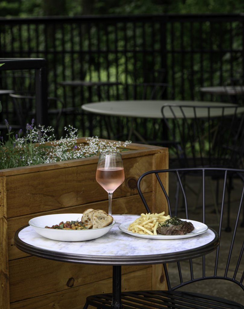 Celebrate Patio Season at Some of Franklin's Best Outdoor Dining Experiences A romantic outdoor dining setup with a glass of rosé wine, a plate of steak and fries, and a bowl of salad on a small round table, surrounded by lush greenery and blooming flowers in Williamson County, Tennessee.