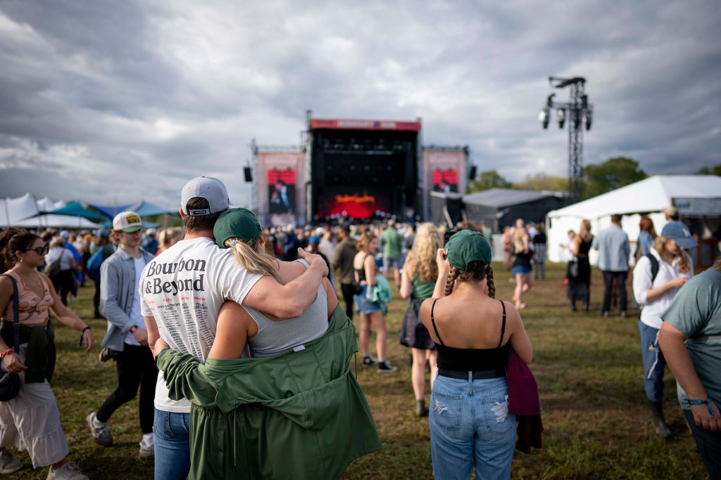 A couple wearing matching green jackets and hats embrace at an outdoor music festival, with a large crowd and stage in the background in Williamson County, Tennessee.