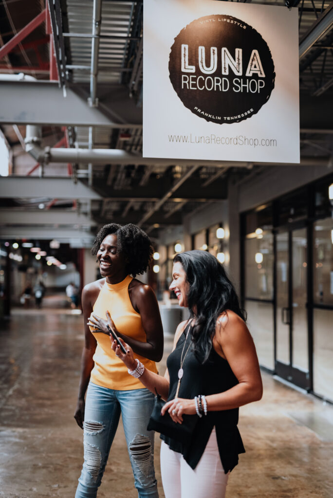 2023-Official-Vacation-Guide-DIGITAL-1 Two women in casual attire walk through a modern indoor space with a record shop sign overhead in Williamson County, Tennessee.