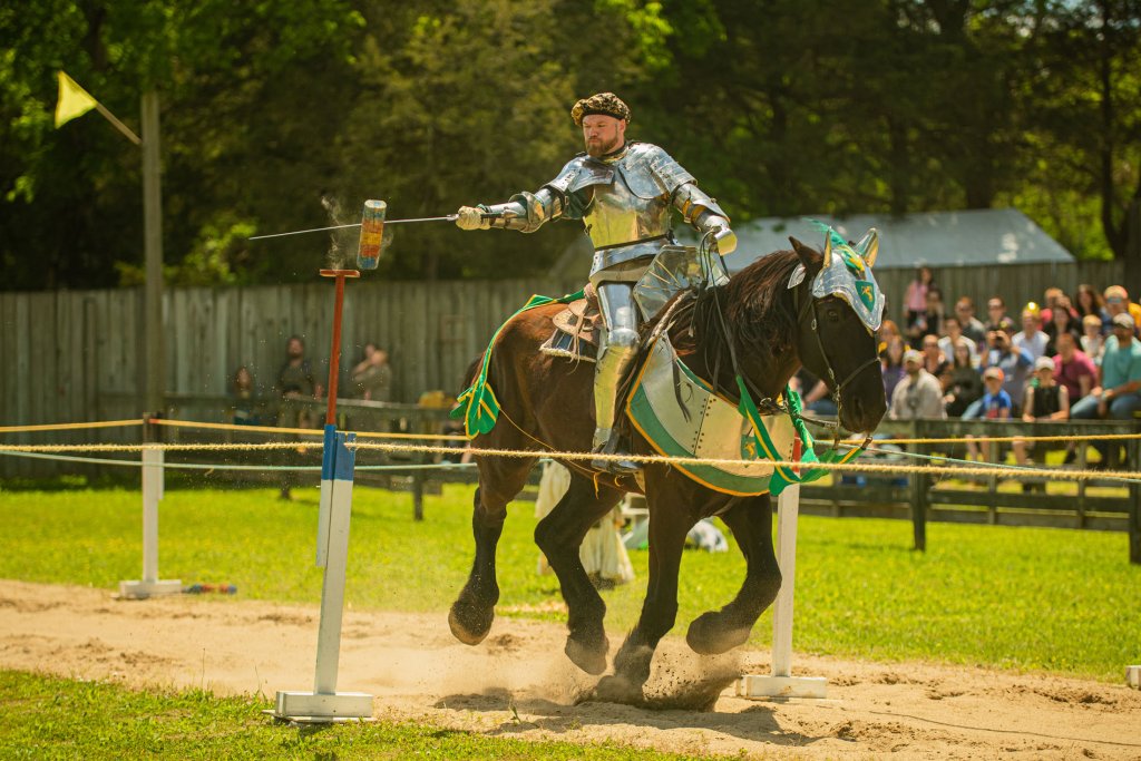 A knight in full armor charges on horseback, brandishing a lance at a target, while spectators watch from behind a fence in Williamson County, Tennessee.