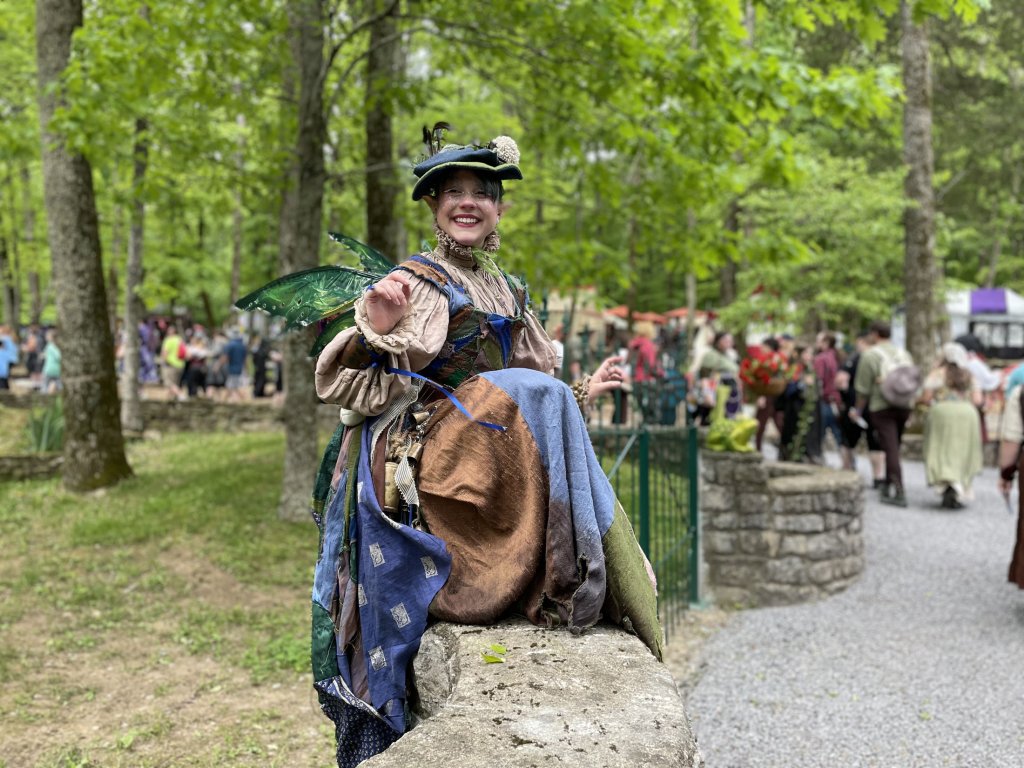 A woman dressed in a whimsical fairy costume with green wings and a hat sits smiling on a stone wall in a lush forest setting, surrounded by a crowd of people in period attire in Williamson County, Tennessee.