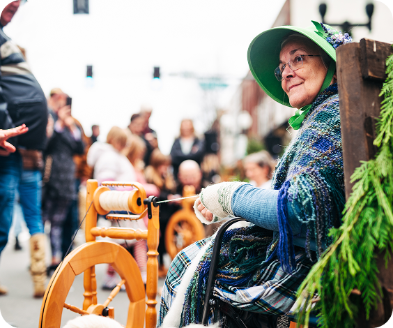 Dickens of a Christmas Elderly woman in traditional attire spins yarn on a wooden wheel amidst a crowd in Williamson County, Tennessee.